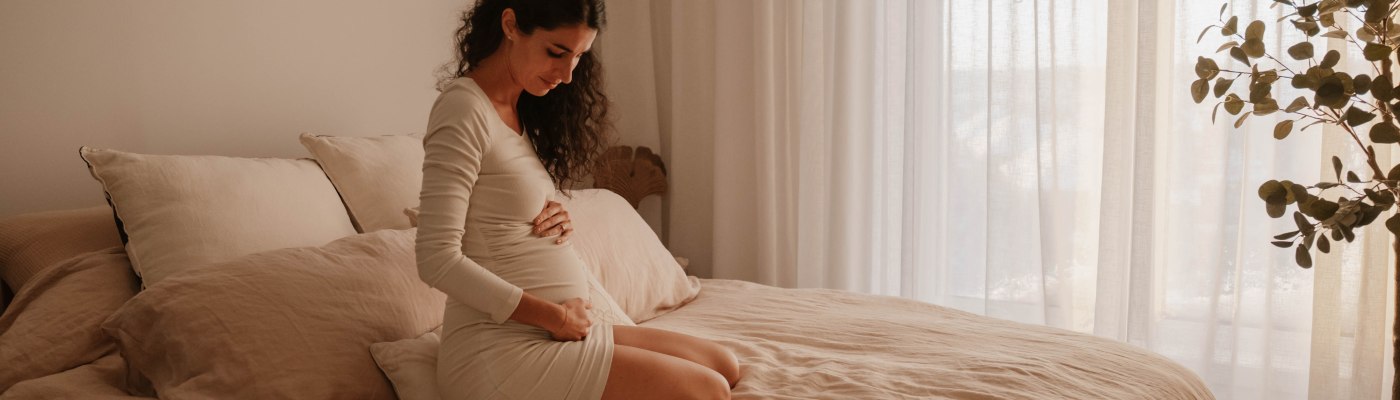 Femme dans ses pensées et assise sur son lit en draps de lin tenant dans ses mains son bedon tout rond de grossesse